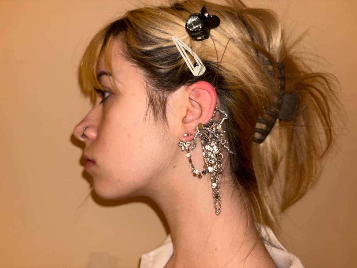 Side profile view of a person with blonde hair styled up, wearing an elaborate silver ear cuff with multiple charms, chains and decorative elements cascading from the ear
