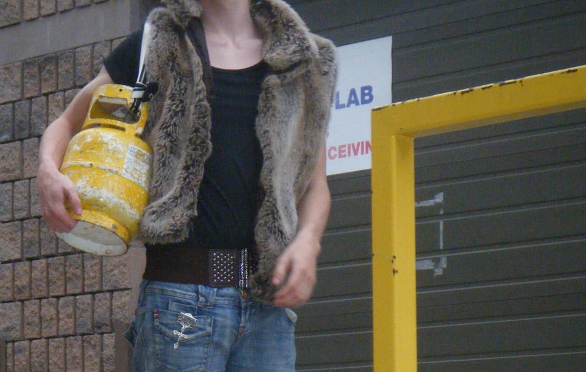Person in fur vest holding yellow propane tank standing in front of industrial doorway with "LAB" sign visible