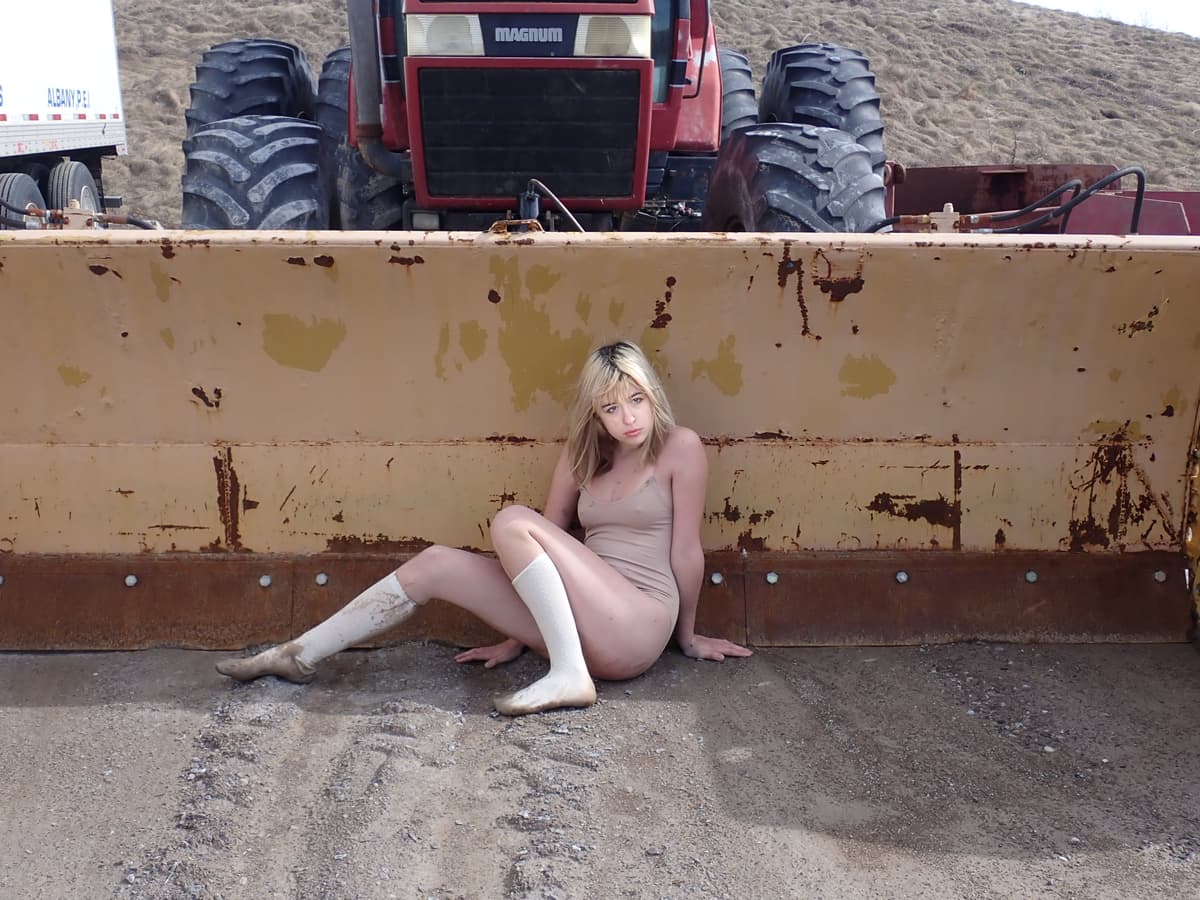 Person in a beige bodysuit sitting against a rusty yellow bulldozer blade with a red tractor visible in the background, wearing white knee-high socks