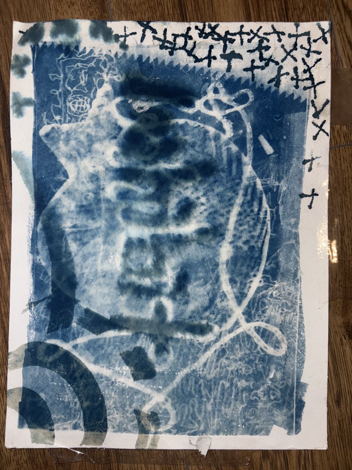 Abstract cyanotype print with white ghostly shapes and patterns against a blue background, featuring cross-hatched markings along the border and circular elements.