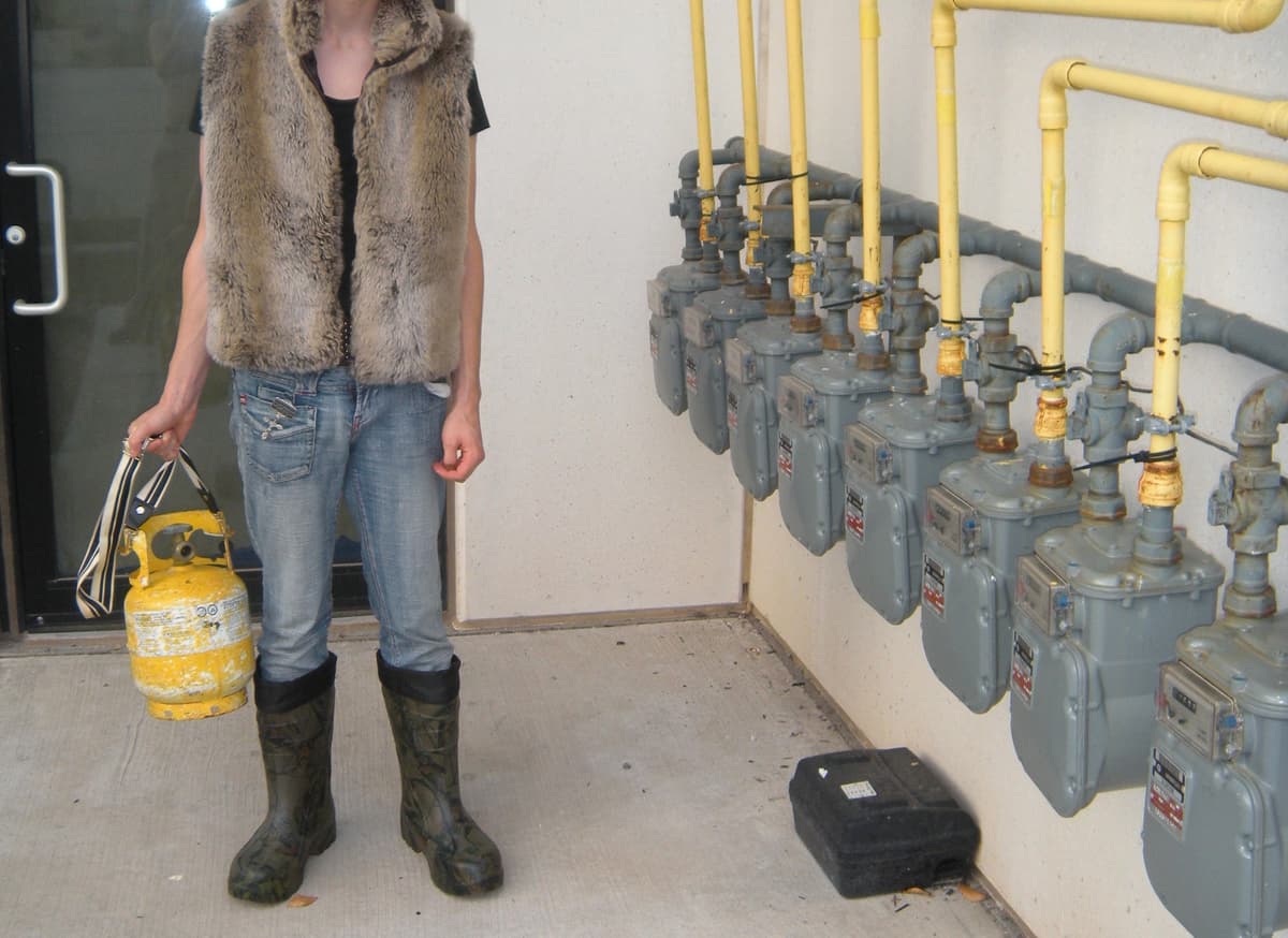 Person wearing fur vest and camouflage boots in utility room with gas meters and yellow pipes mounted on wall, holding yellow propane tank
