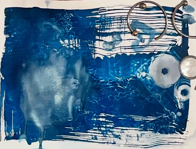 Abstract cyanotype print in deep blue tones with white streaks and patterns, featuring metallic circular elements on the right side against a textured background.
