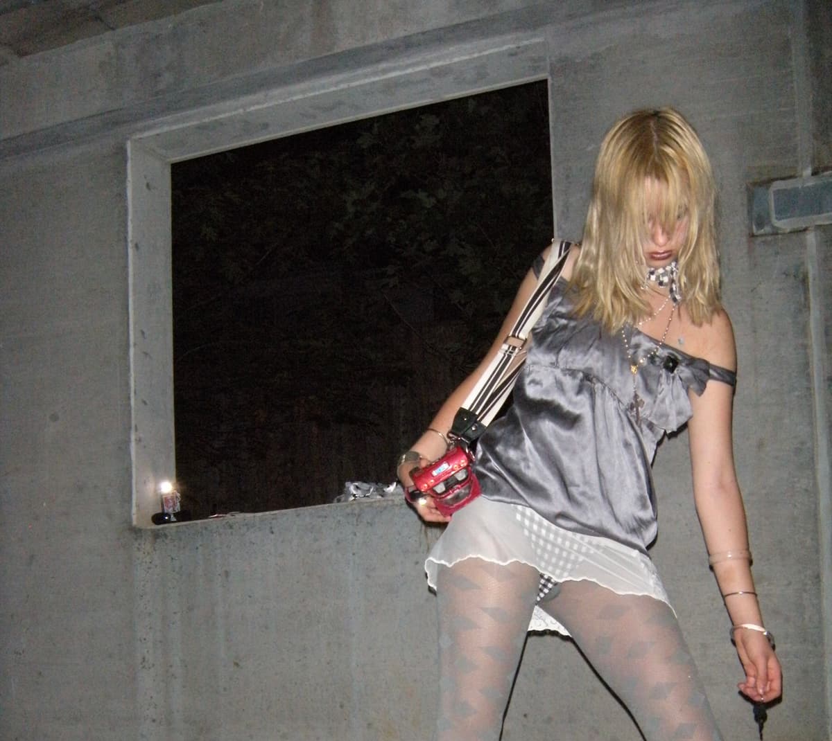 Person with blonde hair wearing silver top and white skirt with gray tights against concrete wall, holding camera with red component
