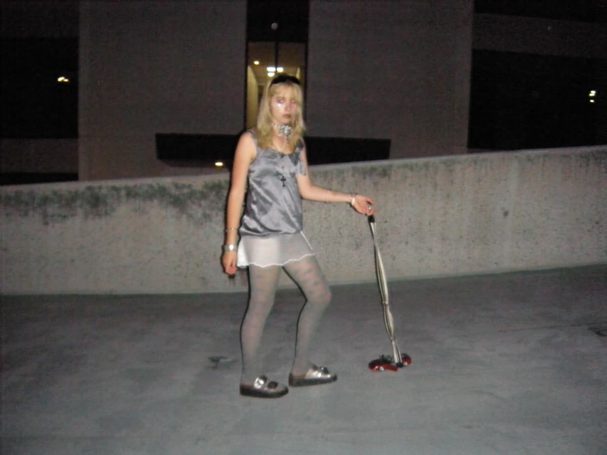 Person in silver outfit and white skirt standing on concrete surface at night holding camera tripod with red component, building visible in background