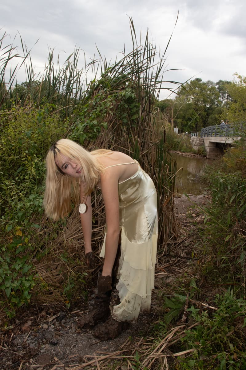 Person in a cream silk dress bending down among tall reeds and wetland vegetation near a small stream, with a footbridge visible in the background