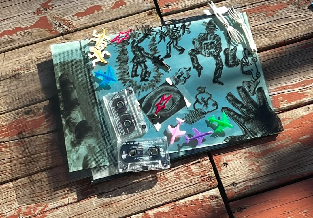 Mixed media assemblage photographed on weathered wooden planks, featuring cassette tapes, colorful plastic toy airplanes, and cyanotype prints with illustrated figures, combining nostalgic objects in an artistic arrangement.