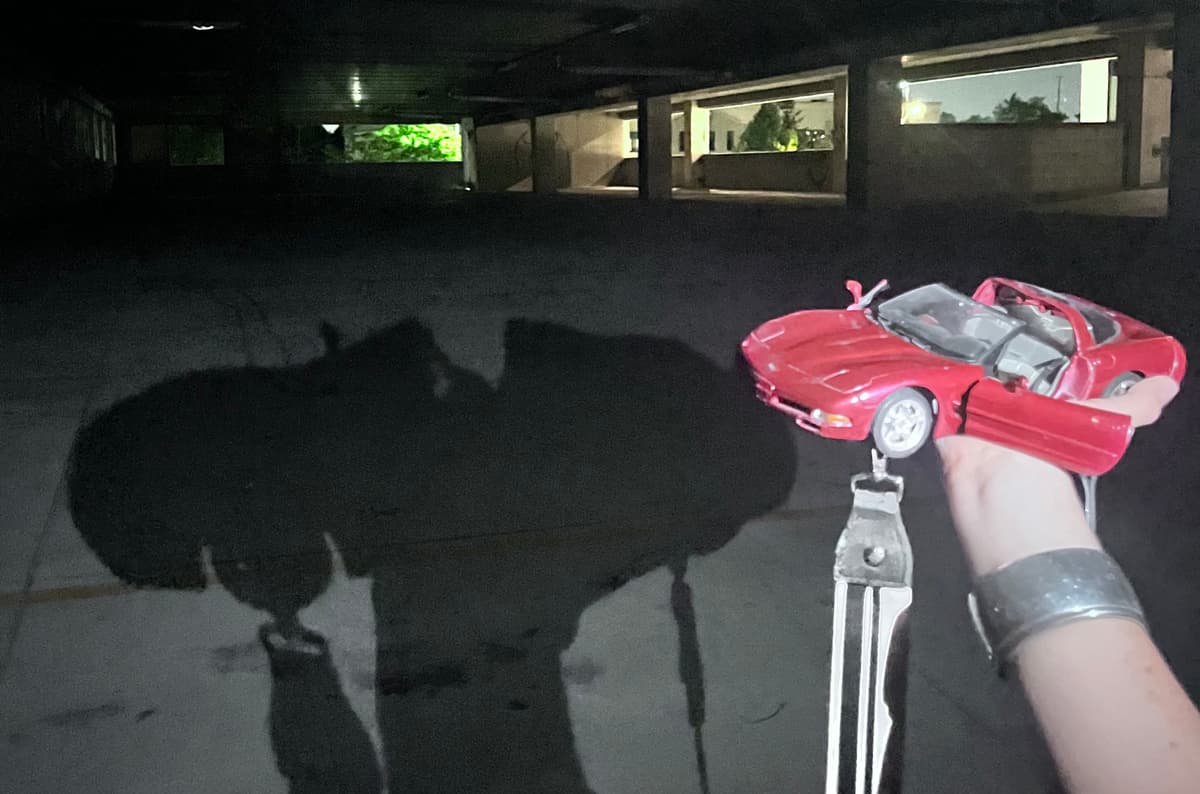 Hand holding small red model convertible car in dark space with shadow cast on floor and windows visible in background