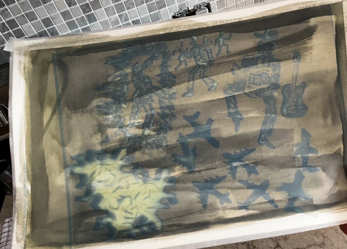 Printmaking screen or stencil showing blue silhouettes of airplanes, human figures, and abstract shapes on a beige background, captured in process on a printing tray.