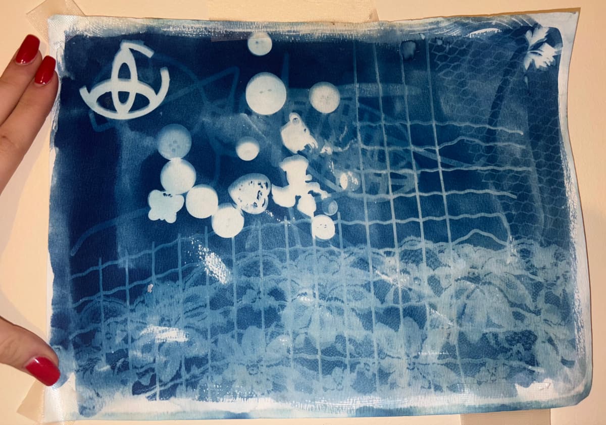 Cyanotype print featuring white grid patterns and circular elements against a deep blue background, with an abstract symbol in the upper left corner and textured wave-like patterns.