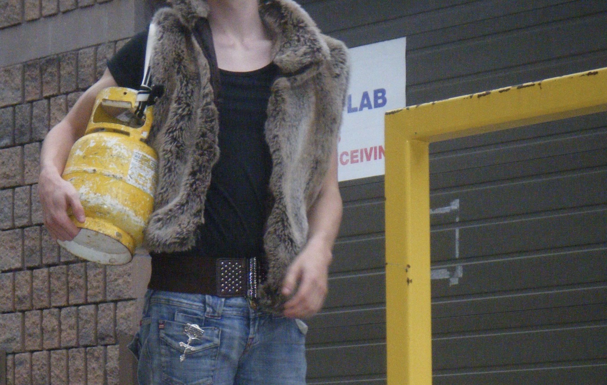 Person in fur vest holding yellow propane tank standing in front of industrial doorway with "LAB" sign visible
