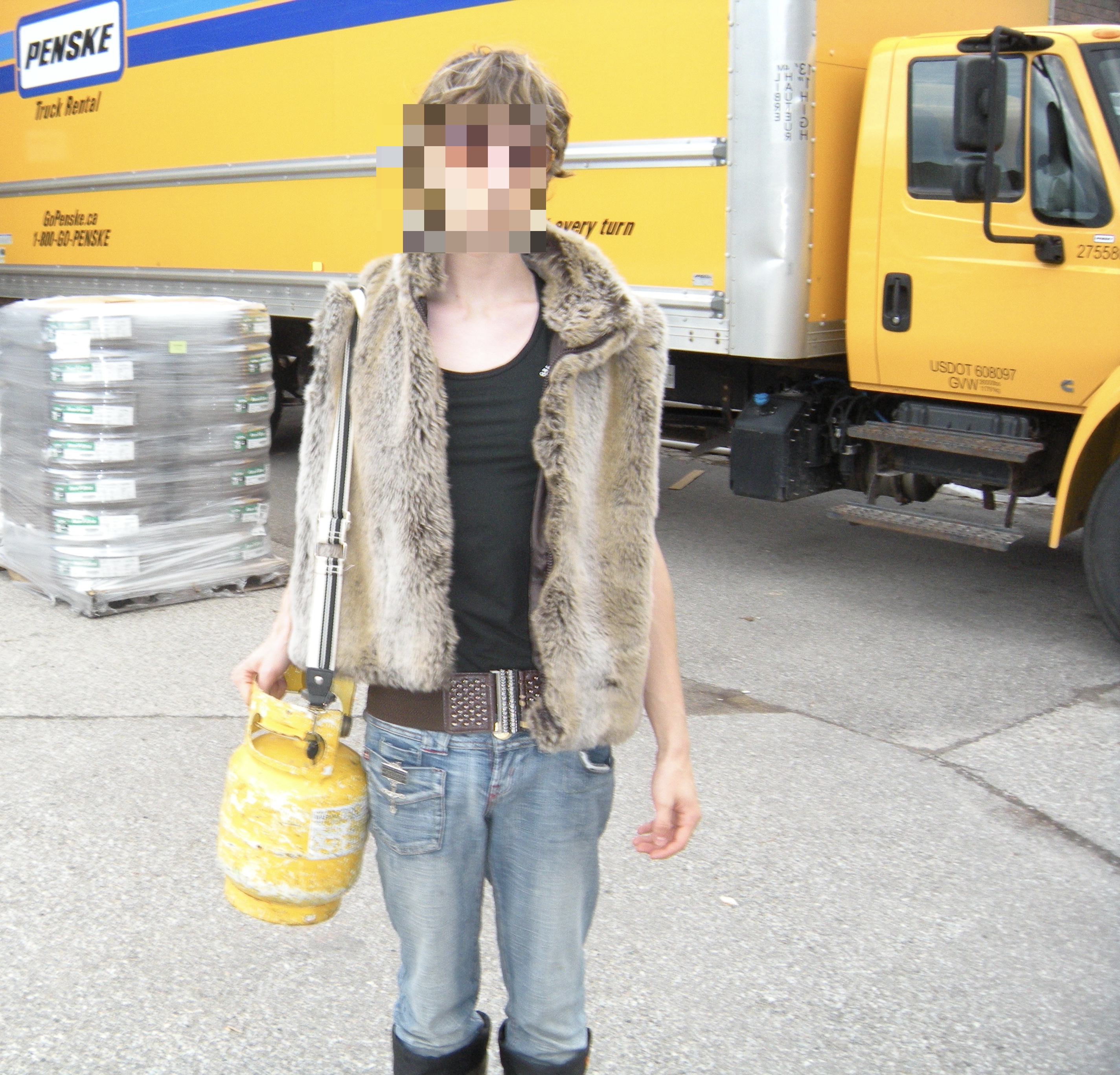 Person in fur vest and jeans holding yellow propane tank standing in front of yellow Penske truck with pallets of packaged goods nearby