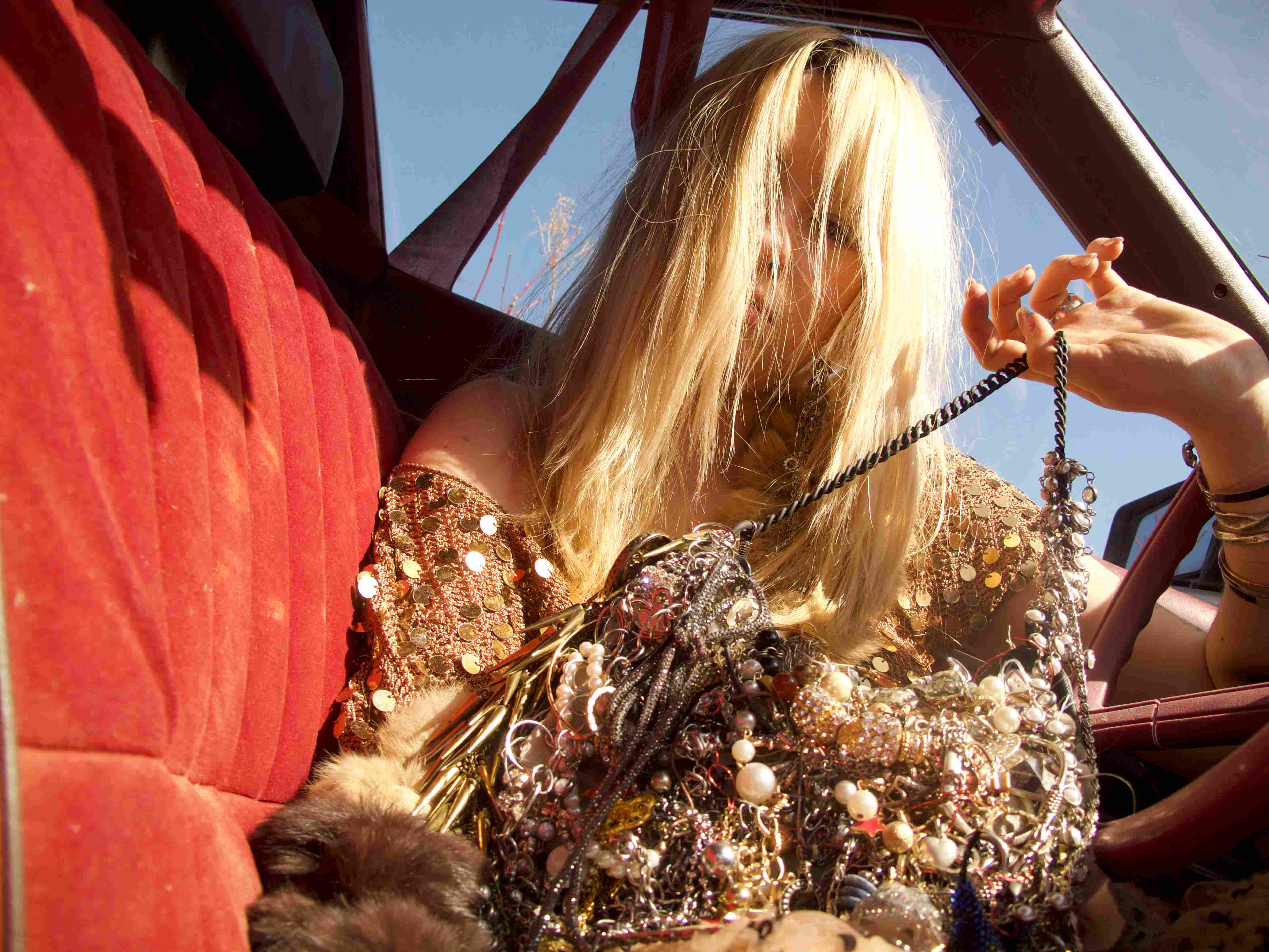 Blonde woman in sequined embellished top sitting in red convertible car interior, holding small furry animal, wearing ornate jewelry with dramatic styling and golden hour lighting