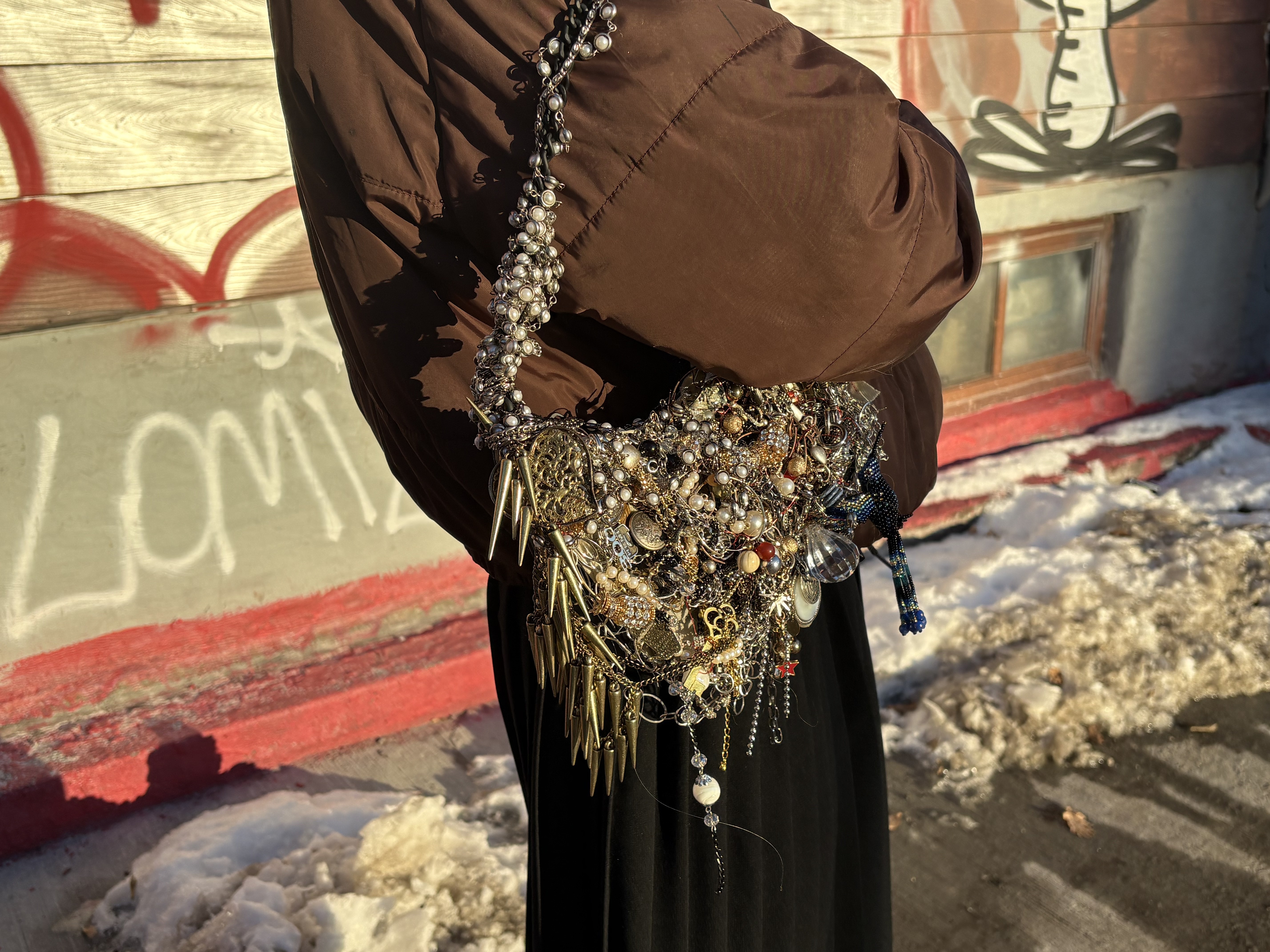 Person wearing a brown jacket and black skirt with an elaborate statement necklace featuring gold and silver charms, pearls, and decorative elements against a graffiti background