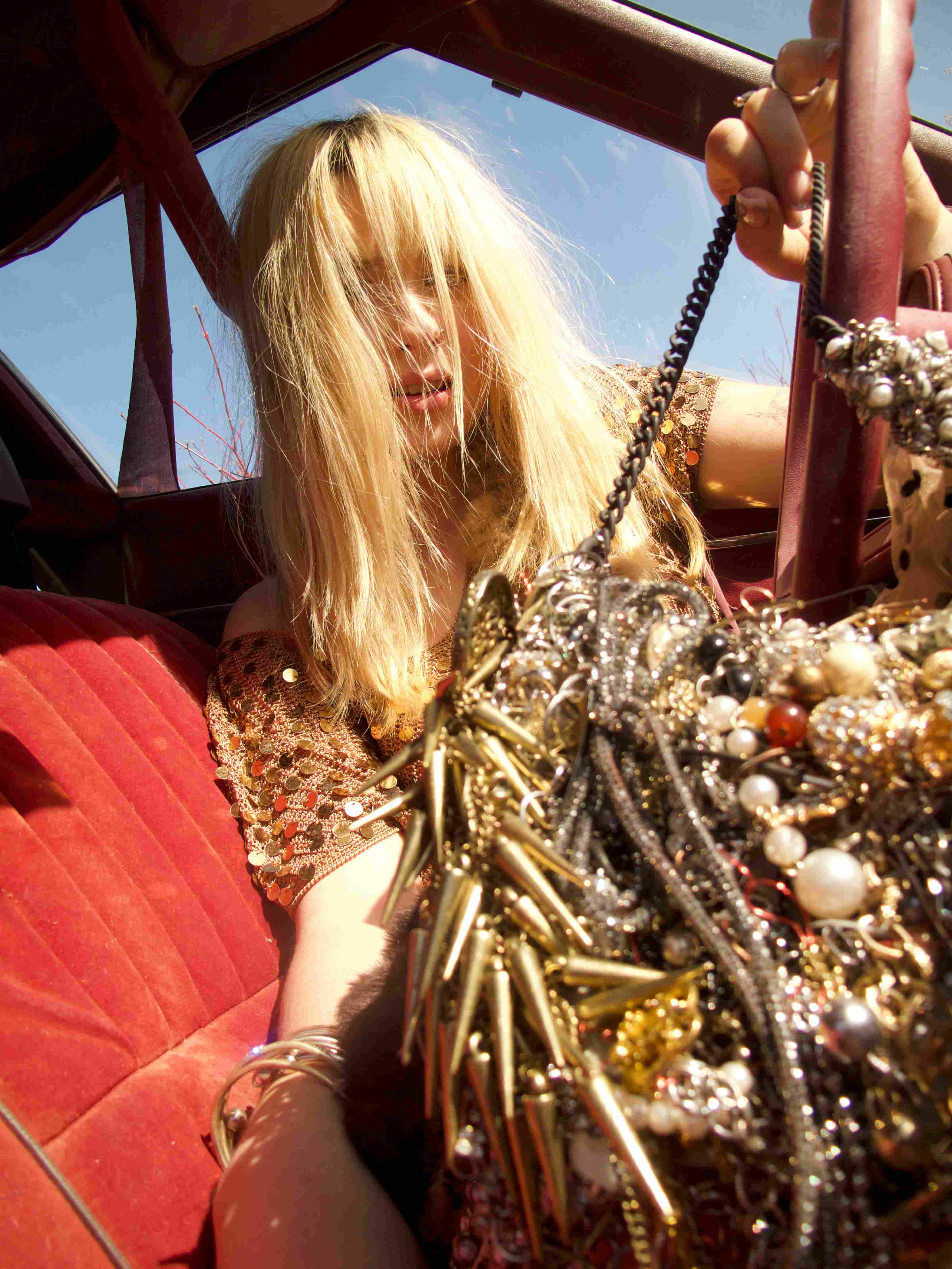 Blonde woman in elaborate sequined outfit sitting in red convertible with top down, wearing extensive metallic jewelry and accessories, captured from side angle with natural lighting