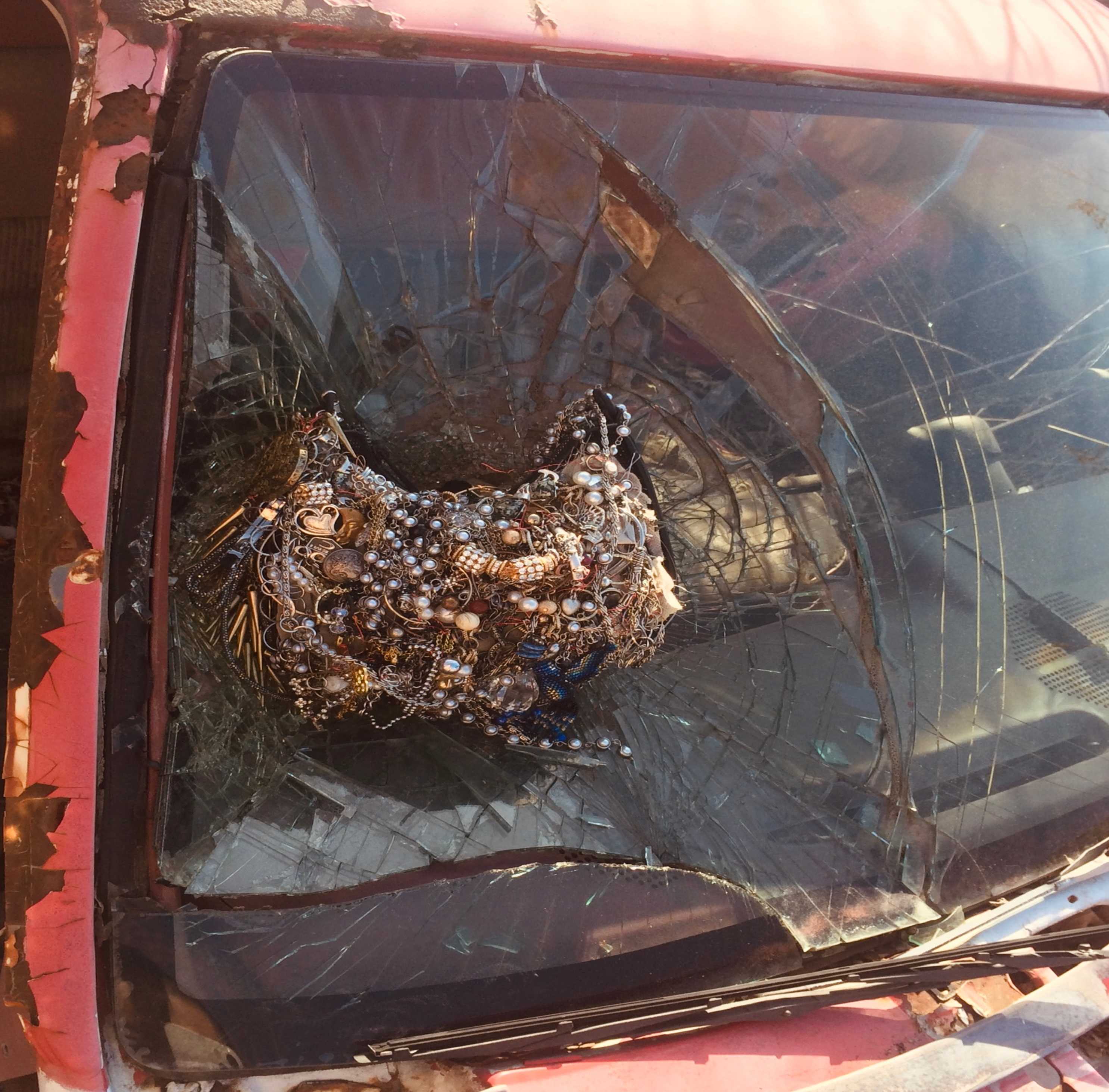 Ornate statement jewelry piece with metallic chains, pearls, and decorative elements displayed on cracked glass surface of car windshield, creating artistic contrast between luxury accessories and damaged glass
