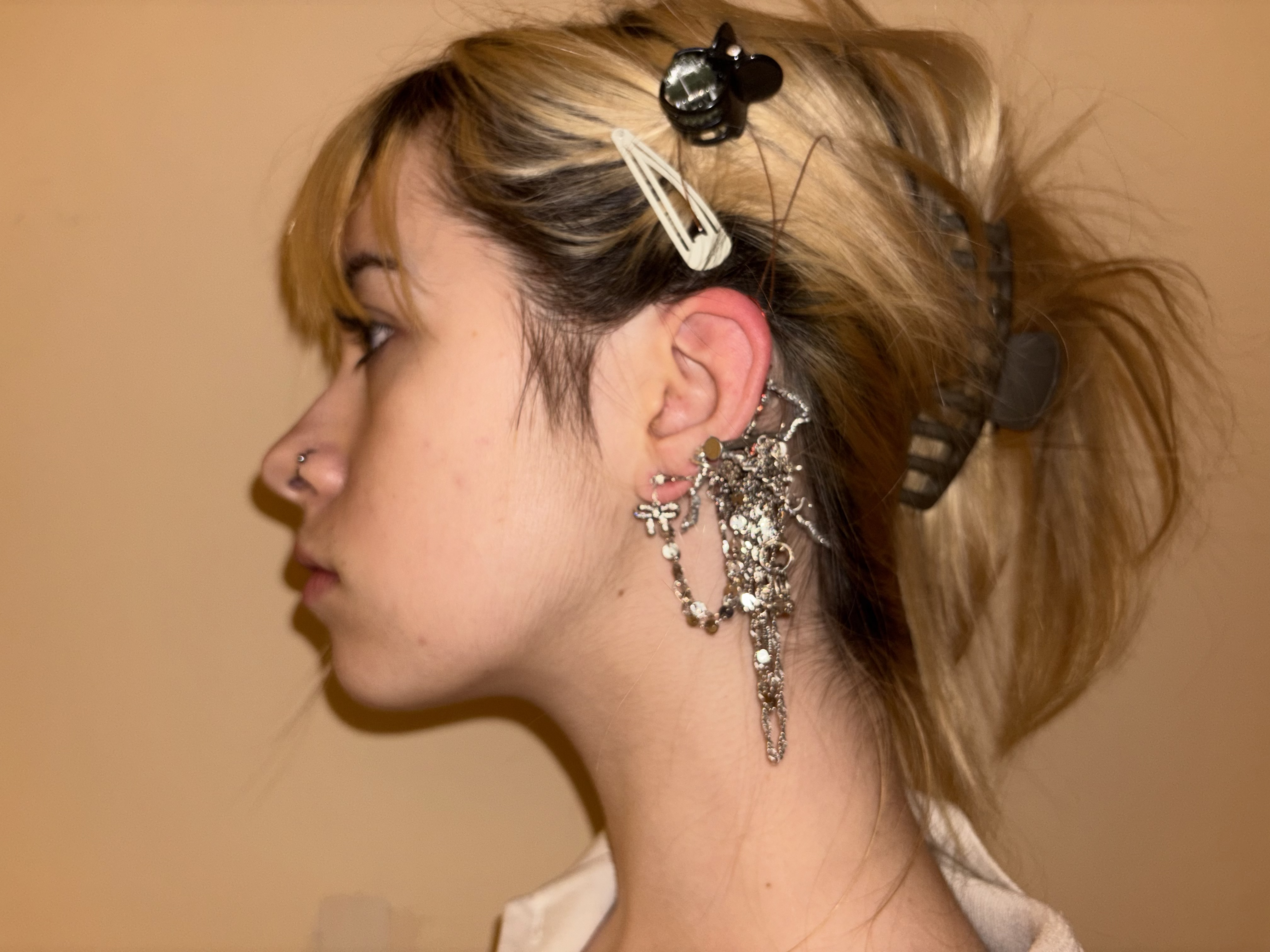 Side profile view of a person with blonde hair styled up, wearing an elaborate silver ear cuff with multiple charms, chains and decorative elements cascading from the ear