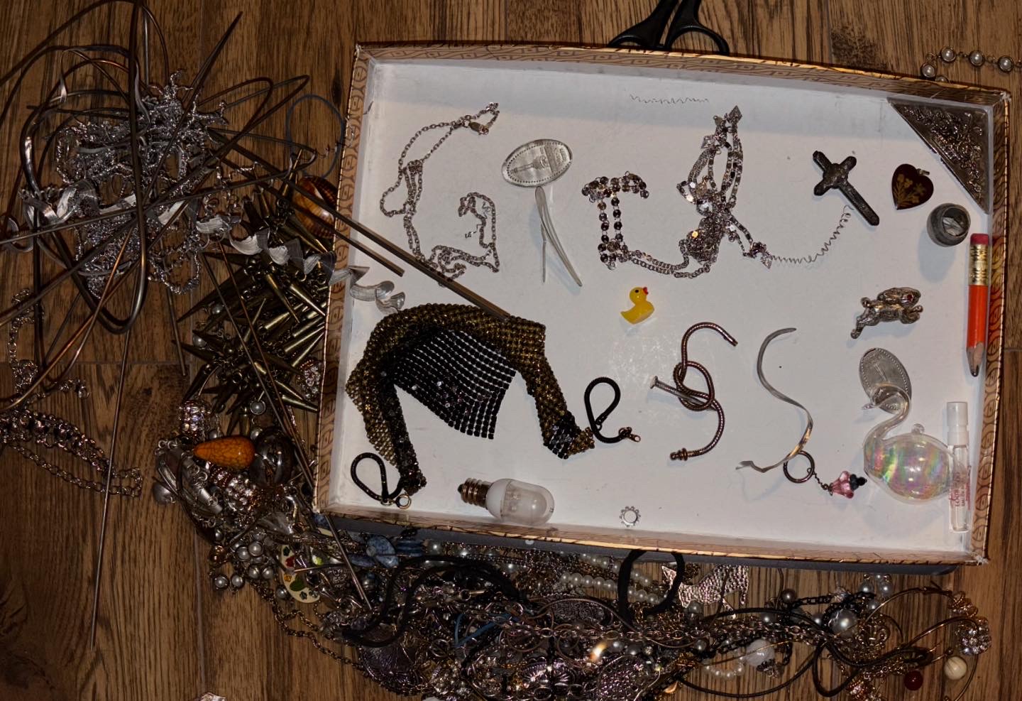 Artistic arrangement of metallic jewelry and found objects in a decorative tray, featuring silver chains, necklaces, a miniature rubber duck, light bulb, and various trinkets displayed on a white surface against wooden background