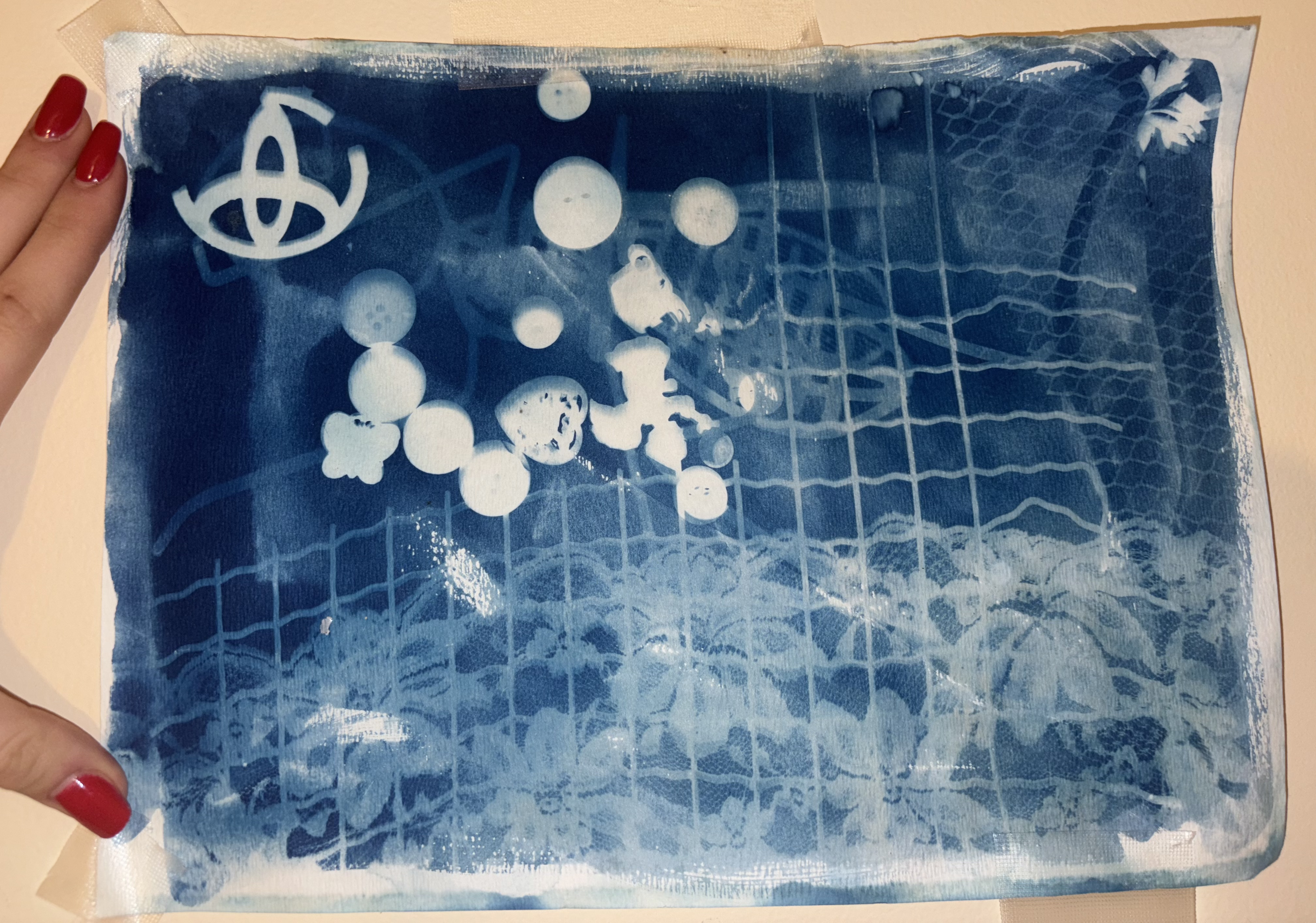 Cyanotype print featuring white grid patterns and circular elements against a deep blue background, with an abstract symbol in the upper left corner and textured wave-like patterns.