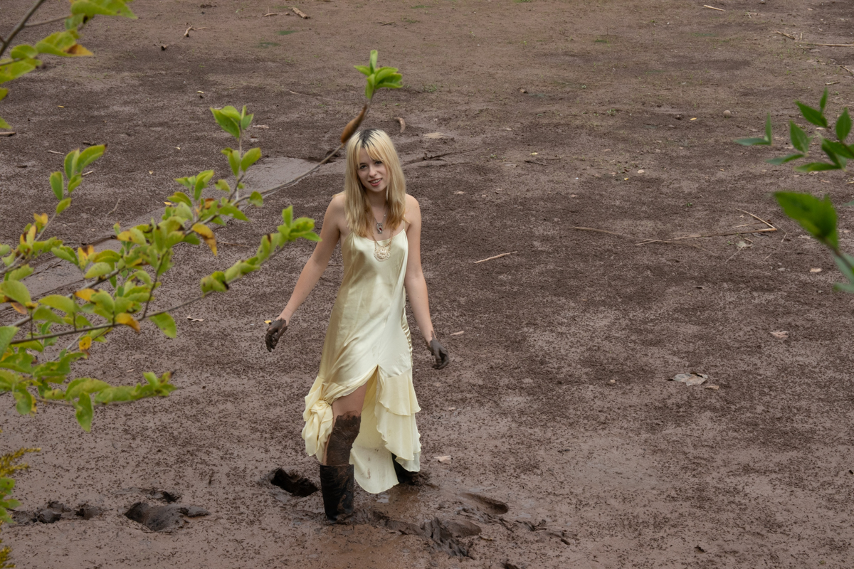 Person in a pale yellow dress and muddy boots standing on wet soil, framed by green foliage with a natural earthy background