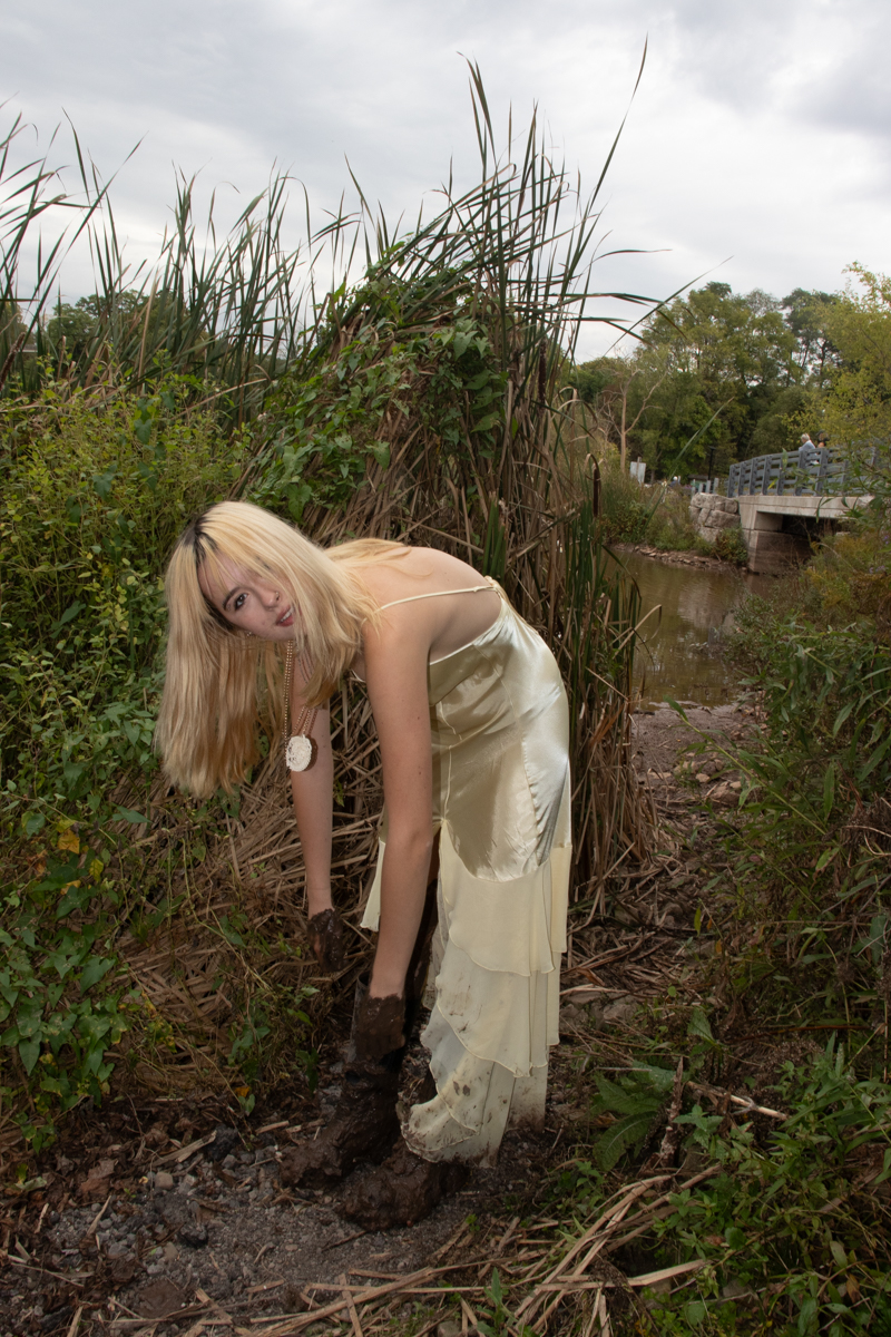 Person in a cream silk dress bending down among tall reeds and wetland vegetation near a small stream, with a footbridge visible in the background