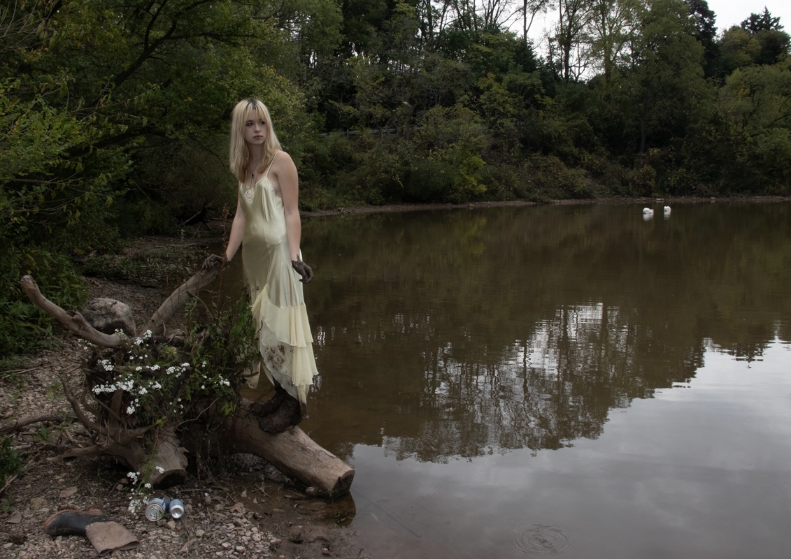 Person in a cream slip dress standing on a fallen log by a calm pond, surrounded by trees in a natural setting with reflections in the water