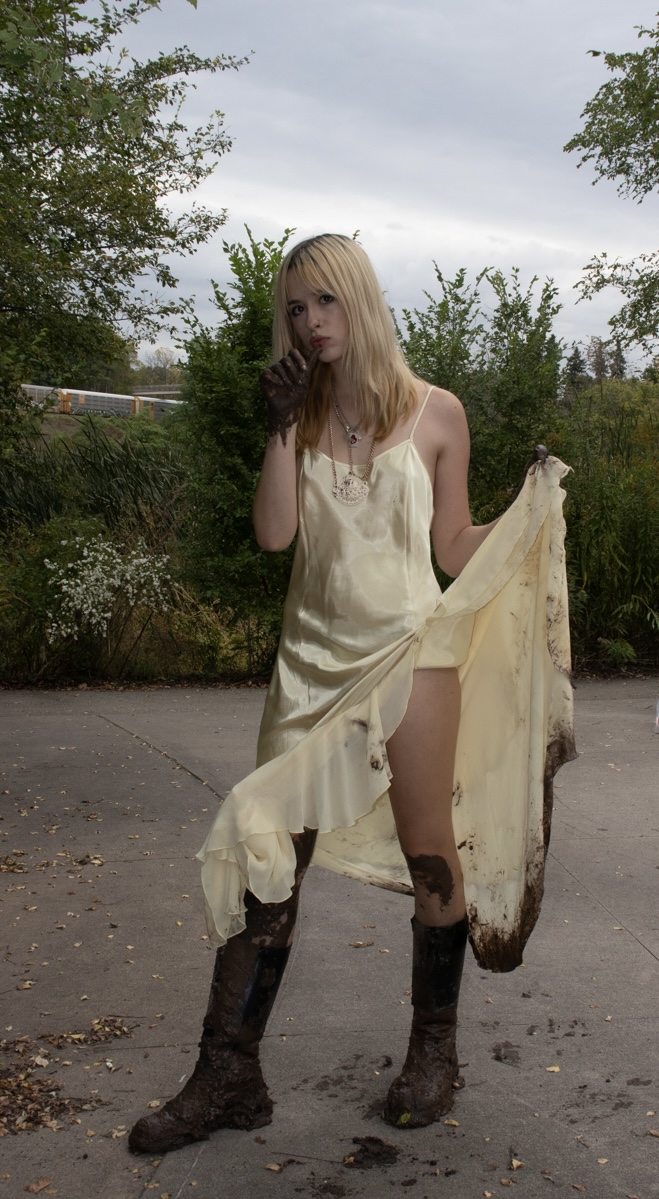 Person in a light-colored slip dress and muddy boots standing on a concrete path, holding part of the dress fabric, with greenery in the background