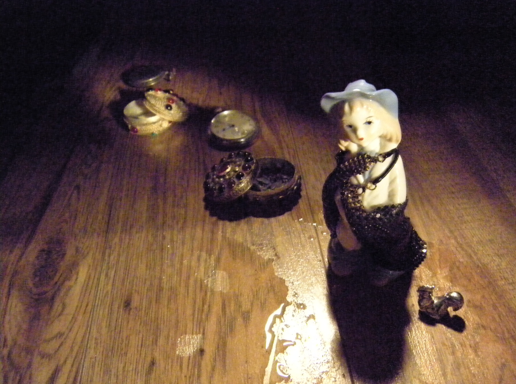 Moody still life photograph of porcelain figurines and antique pocket watches on wooden surface, dramatically lit to create strong shadows and highlight object details