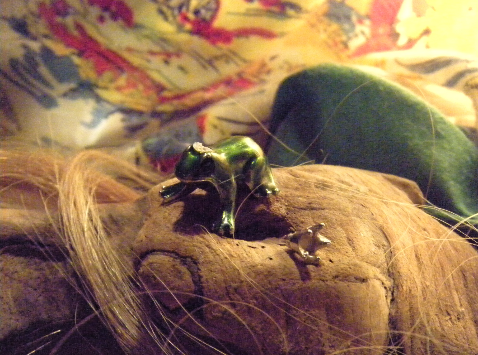 Macro photograph of a metallic green frog figurine perched on textured natural material, with strands of blonde hair and colorful fabric visible in the background