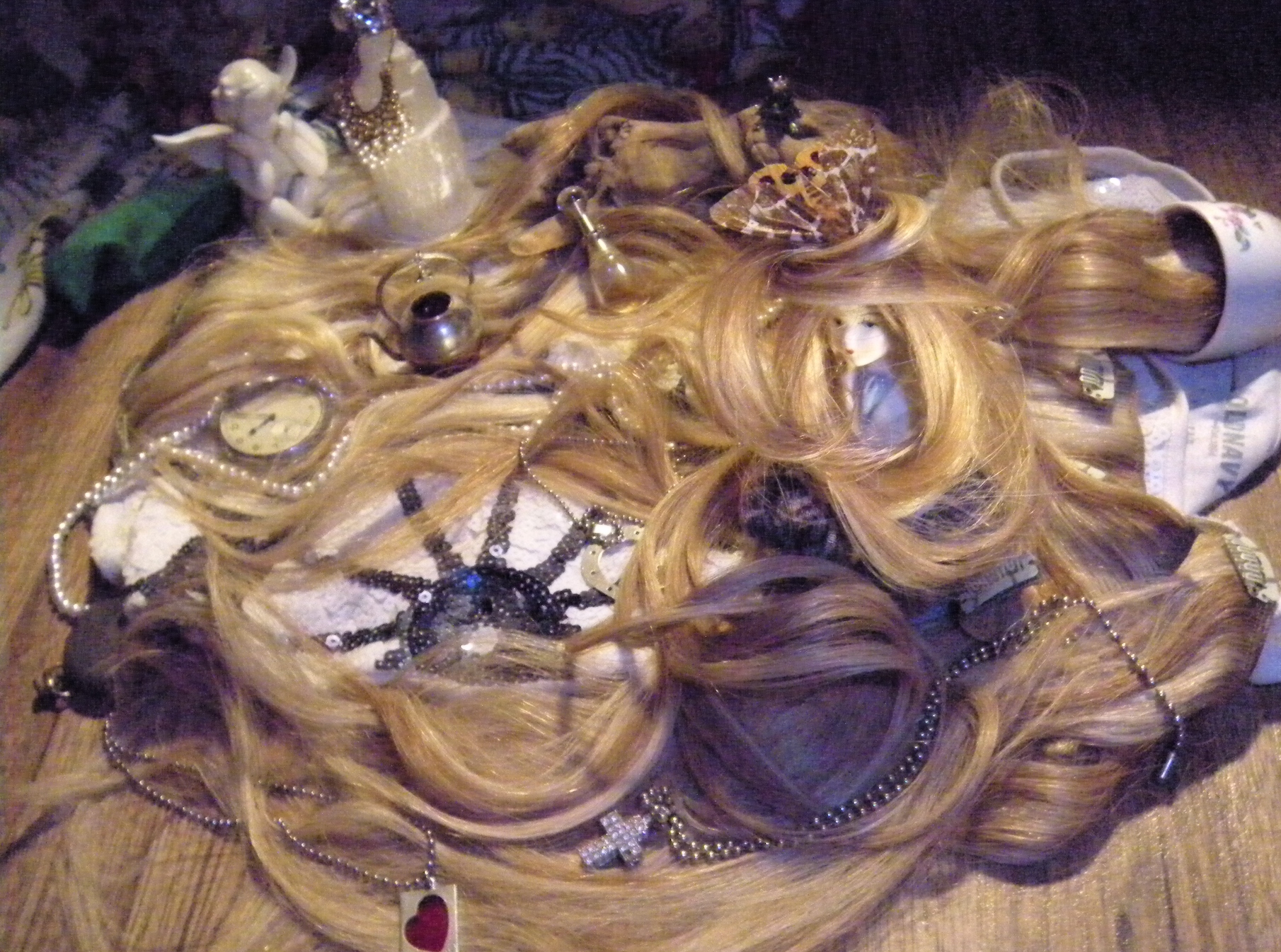 Close-up of blonde hair extensions arranged with embedded decorative elements including beaded crosses, heart-shaped pendants, and vintage trinkets, photographed against wood texture