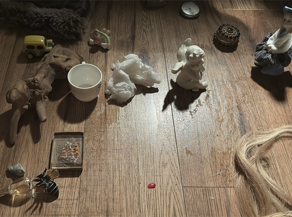 Overhead view of a curated collection of objects on wooden flooring, featuring a yellow toy car, white ceramic cup, tissue paper, porcelain figurines, and various decorative items arranged in a still life composition